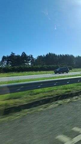 This media file captures a view from a moving vehicle on a sunny day in Rebala, Estonia. The scene is dominated by a bright blue sky with a few wispy clouds. In the foreground, a silver metal guardrail lines the side of the road, with green grass and some darker vegetation visible on either side. Beyond the guardrail, a paved road stretches into the distance, with a line of trees forming a dense forest along the horizon.

The activity taking place is travel by car. Two vehicles are visible on the road: a dark-colored van or SUV passes by in the middle distance, followed shortly by a white station wagon. The sun appears to be high in the sky, suggesting it is daytime, likely late morning or afternoon. The clear blue sky and bright sunlight indicate fair weather conditions. The landscape appears rural, with the prominent forest suggesting a natural or semi-natural environment. No text is visible in the media file. The overall mood conveyed is one of peaceful travel through a scenic, sunlit landscape.