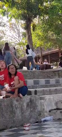 The video shows a group of people sitting on concrete steps outdoors, with lush green trees and foliage in the background. The lighting suggests it is daytime, possibly late afternoon, with sunlight filtering through the leaves. Several individuals are seated on the steps, some facing forward, others in profile.

In the foreground, two women in red shirts are sitting on lower steps. The woman on the left is holding a white cup and appears to be eating. The woman to her right is wearing glasses and is also holding a cup.

Further up the steps, a woman in a light grey hijab and a woman in a dark hijab are visible. Other individuals are scattered across the steps, some of them talking or looking around. A man in a red shirt is standing further back, near a white pole.

In the background, there's a traditional-looking structure with a thatched roof. The overall atmosphere seems relaxed, possibly a gathering or a break in an outdoor activity.

As the video progresses, the perspective shifts, showing more of the environment. A man in a dark blue shirt and dark pants is sitting with his feet in a pool of water, looking and laughing towards the camera. Another person's legs are visible in the foreground of this shot, also near the water. Further back, a woman in a black garment and a face veil sits on the steps, and another person in a black shirt with white lettering is visible, also seated. The video ends with a focus on the man with his feet in the water.