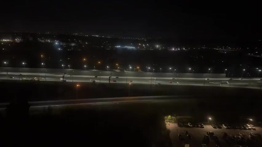 A nighttime aerial shot shows a highway with traffic and a parking lot. The highway is illuminated by streetlights and the headlights and taillights of vehicles. Cars and trucks are visible on the highway, moving in both directions. A large parking lot with many cars is visible in the lower right corner of the frame. The background is dark, with some distant lights suggesting a city or town.
