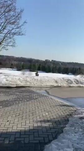 The video shows a view of a park or landscape on a sunny day with patches of snow still present. The camera pans slowly from left to right, focusing on a brick or stone-paved path in the foreground. Patches of snow are visible on either side of the path, gradually receding. Beyond the path, there is a landscape with trees and grass, some of which is still covered in snow. In the distance, there are buildings, suggesting a residential or urban area. The sky is clear and blue. There are no people or animals visible in the video.