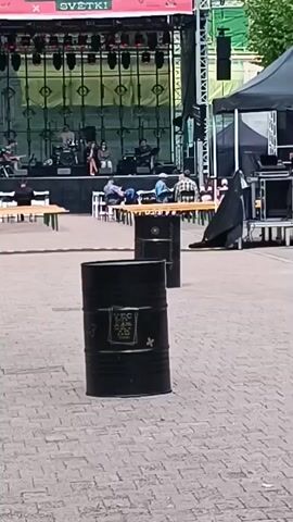 The video shows an outdoor setting with a stage in the background. On the stage, a band is performing, with several musicians visible playing instruments. In the foreground, there are three black barrels arranged in a line. The ground is paved with cobblestones. There are picnic tables and chairs set up in front of the stage. A black tent is also visible to the right of the stage. The overall scene appears to be a public event or festival.