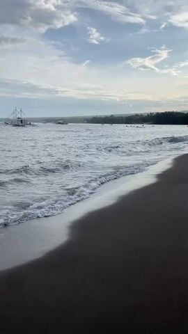 The video shows a beach with dark sand meeting the ocean. Gentle waves are rolling onto the shore. In the distance, several boats are visible on the water, with one larger white boat in the foreground left. There are people in the water, appearing to be swimming or wading. The sky is cloudy with patches of blue, suggesting it is either morning or late afternoon. A treeline is visible along the horizon.