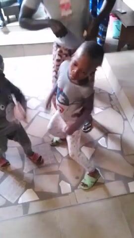 A young child wearing a gray shirt, patterned pants, and green sandals dances to music on a tiled floor. Other people, including another child, are visible around the dancing child, and money is briefly thrown in the dancer’s direction. The setting appears to be indoors, with a wooden staircase and other furniture visible in the background.