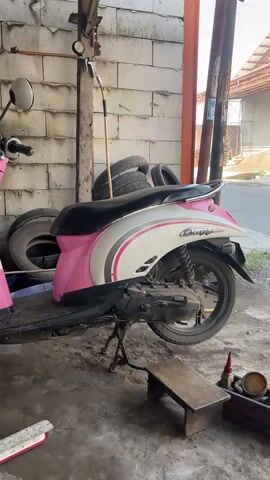 The video shows a Honda Scoopy scooter propped up on its kickstand. The scooter is pink, white, and black, and it appears to be in need of maintenance, judging by the dirt and grease on the engine and wheel. A small wooden stool sits next to the rear tire, and a toolbox with various items rests beside it. In the background, there is a building with stacked tires visible, and a person on a scooter drives by on the street. The scene appears to be a repair shop or garage setting.