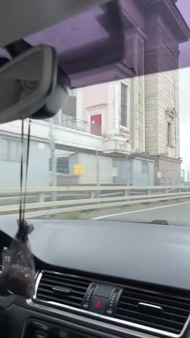 The video is shot from inside a car, looking forward through the windshield and to the right. The driver's hands are visible on the steering wheel. A car air freshener hangs from the rearview mirror. The car is moving on a road with a fence and buildings on the right side. In the distance, there is a body of water with trees on the far bank. The sky is overcast.