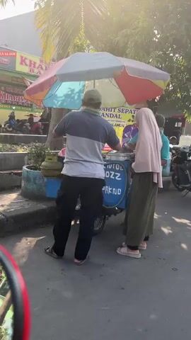 The video depicts an outdoor scene with a food cart. A man wearing a grey and blue shirt, a green cap, and black pants stands behind the food cart, appearing to be serving customers. In front of him, two individuals are visible. One is a woman wearing a light brown hijab and a long green skirt. The other person is partially obscured but appears to be wearing a blue garment. The food cart is adorned with a colorful umbrella and has text on its side, though it is difficult to read clearly. A gas cylinder is attached to the cart. The background shows buildings, a street, and some parked motorcycles. Sunlight is prominent, casting shadows and creating a bright atmosphere.