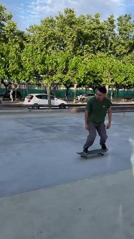 A young man is skateboarding at an outdoor skate park in Santa Perpètua de Mogoda, Spain. The park features a concrete surface with various obstacles, including a low metal rail in the center of the frame and a small concrete bank or quarter pipe on the left. In the background, numerous green trees line the area, with cars parked on a street and buildings visible further in the distance, indicating an urban or suburban setting. The sky is clear and bright with some white clouds, suggesting it's daytime, possibly late afternoon.

The skater, wearing a green t-shirt, grey cargo pants, and black sneakers, begins by rolling on his skateboard. He then approaches the metal rail and successfully performs an ollie, jumping with his skateboard over the rail and landing smoothly on the other side. A wooden structure behind the rail has the word "LIBERA" painted on it.
