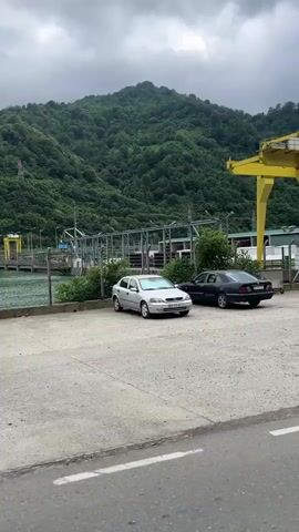 A panoramic video pans across an industrial area with a large yellow gantry crane dominating the landscape. In the foreground, a dark-colored car is parked in a gravel lot. Behind the car, a low-lying building with a grey roof and concrete base stands. Further back, a large white building with a metal exterior is visible, adjacent to a chain-link fence. Beyond this, the terrain slopes upwards, covered in dense green forest. Another yellow gantry crane is visible further in the distance. The sky is overcast and grey. The video appears to be taken from a vehicle moving along a paved road.