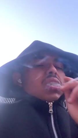 The video features a person in a dark hooded sweatshirt taking a selfie-style video. Initially, the camera is pointed upwards, showing the person's face partially obscured by the hood and a bright sky in the background. A close-up shows a white earbud (likely an AirPod) in their ear. The scene then shifts to another young man with dark hair, wearing a red and white varsity-style jacket and light pants. He is sitting and looking at his phone. Next to him is a white LG air conditioning unit with a green can on top. The video concludes with a view of a red-tiled rooftop, a metal chimney with a black cap, and the top of a white outdoor air conditioning unit, on which a small red and white box and another dark object are visible. The time of day is daytime with a clear sky, suggesting an outdoor or semi-outdoor location, possibly a balcony or rooftop in Amersfoort, Netherlands.