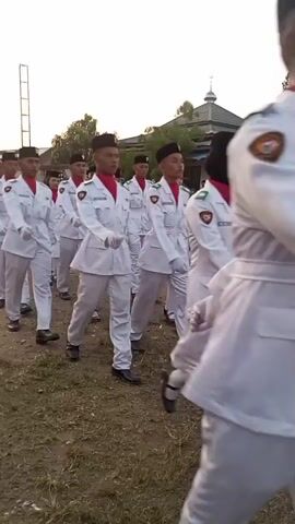A group of young people in white uniforms with red scarves and black hats are marching outdoors in a line, with a mosque in the background. Some of the marchers are looking straight ahead, while others are glancing to the side. In the background, there are spectators and some trees. The ground is covered in dry grass and dirt. The lighting suggests it is either early morning or late afternoon.