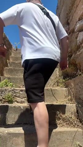 The video shows a man walking up a set of stone steps. He's wearing a white t-shirt, black shorts, and black Nike shoes. He has a black Hugo Boss bag slung across his shoulder. He is bald. The steps are old and weathered, with some vegetation growing nearby. Behind the man is a clear blue sky. At the top of the stairs, a stone wall and some sparse vegetation are visible. The man pauses and looks toward the camera.