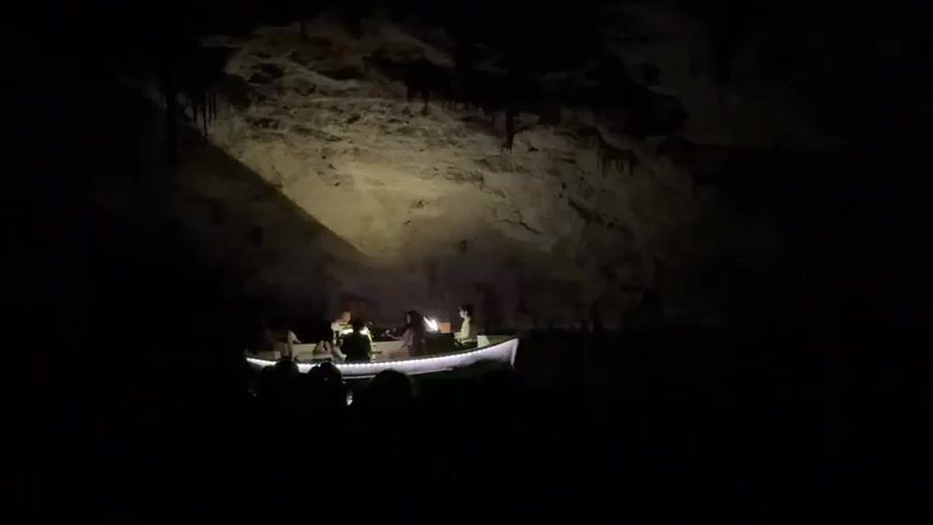 In a dimly lit cave, a small rowboat with several people on board is illuminated by spotlights. The boat has a white hull and is lit from below by a string of lights. The people in the boat appear to be musicians, as they are holding instruments. The cave walls are textured and uneven, with some stalactites hanging from the ceiling. There are silhouettes of people watching from the dark foreground. The atmosphere is mysterious and dramatic.