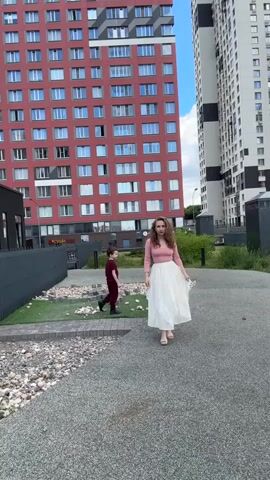 A woman in a pink long-sleeved shirt and a white skirt walks towards the camera on a paved path. Behind her, a young boy in a maroon shirt and pants runs away from the camera. They are in an urban environment with tall, modern buildings behind them. The building on the left is red with many windows, and the building on the right is white and gray with black stripes. The area around the path is covered in small stones and some patches of artificial grass. The sky is cloudy.