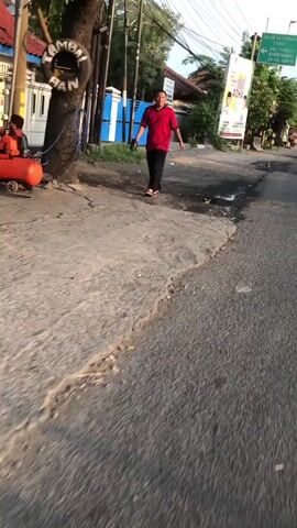 The video shows a man walking on a paved road near a tree. He is wearing a red shirt and black pants. There is a tire hanging from the tree and an orange air compressor on the ground next to it. The road is lined with buildings and vegetation. The sun is shining, casting shadows on the ground.