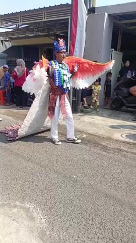 This video shows a street parade. A person in a peacock costume walks down the street, followed by a group of people in matching red outfits. The person in the peacock costume is wearing a large, elaborate costume that includes a feathered headdress, a colorful beaded necklace, and a long, flowing tail that resembles a peacock's feathers. They are also holding a large, fan-like appendage that is red and white. The people in red outfits are wearing headscarves and traditional clothing. One person in the red group is holding a large umbrella that is decorated with flowers. The parade is taking place on a sunny day, and there are buildings and trees in the background. The street is paved, and there are some spectators watching the parade from the side of the road.