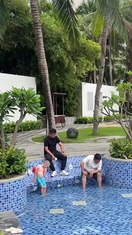 A boy in swim trunks and arm floaties stands at the edge of a tiled pool. Two men sit on a bench partially submerged in the pool. One man is wearing a black shirt and pants. The other man is wearing a white shirt and shorts. The background shows palm trees, bamboo, a white building, and green grass.