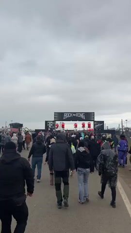 A wide shot of an outdoor event shows a large crowd of people walking towards a stage. The stage has a banner that says "ROCK AM RING" at the top and the letter "T" in pink at the bottom left. There are four large red objects on the stage, possibly speakers or decorations. The people are dressed in casual clothing, and some are wearing hooded jackets or raincoats. The ground is a dirt path, and the sky is overcast. On the left side of the frame, a person is holding a glass of beer.