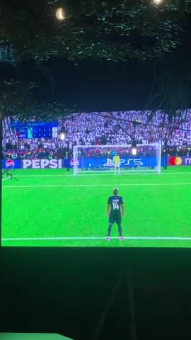 A TV screen shows a soccer player on the field, facing the goal, about to take a penalty kick. The player is wearing a blue uniform with the number 14 on his back. The goalkeeper is in a yellow uniform. The crowd is visible in the background. The penalty score is shown on the screen in the upper left. Advertising for Pepsi, Playstation and Mastercard is also displayed on the screen. The screen is viewed from underneath a tree with hanging lights.