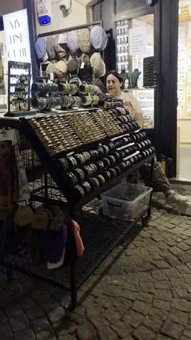 The video appears to capture a bustling street scene at dusk in Urla, Turkey, likely in a commercial area with shops and cafes.

In the foreground of the first scene, a woman with dark hair smiles while standing behind a display of various jewelry items, including necklaces and bracelets. The display stand is well-stocked, suggesting a retail environment.

The subsequent scenes shift to a street lined with businesses, illuminated by artificial lights, indicating it is evening. People are seen dining at outdoor tables of cafes and walking along the cobblestone street. The atmosphere seems lively and social.

One scene shows a man standing in the doorway of a shop. Another scene focuses on a group of people seated at tables outside a cafe. Further down the street, other patrons and pedestrians are visible. Some shop signs are partially discernible. One sign reads "IRMIK HANIM PASTRISSERIE." Another sign, partially obscured, appears to be for "GUVEN GLASSES." A child wearing a blue shirt with "RONALDO 7" on the back is seen in the foreground of one shot.

The time of day is evening, suggested by the artificial lighting and the relaxed atmosphere of people enjoying themselves outdoors. The weather appears to be pleasant, suitable for outdoor dining and strolling. The cobblestone street and the architecture of the buildings suggest a charming, possibly historic, urban setting. The overall mood conveyed is one of a vibrant evening in a Turkish town.