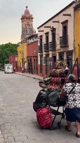 Here is a description of the video:
The video depicts a street in what appears to be a historic Mexican town or city. The street is paved with cobblestones and lined with colorful buildings. The buildings are mostly two-story structures with balconies and wrought iron details. In the background, a church tower is visible with a red dome. A woman is standing on the street next to a cart. The cart is filled with a variety of items, including what looks like wreaths. She appears to be organizing or adjusting something on the cart. There is a white bucket hanging on the side of the cart. To the left, a white vehicle drives down the street. Several people can be seen walking or standing along the street in the background. The sky is overcast, suggesting it is either early morning or late afternoon.
