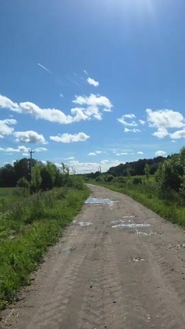 The video shows a dirt road with puddles of water reflecting the blue sky and white clouds. The road is surrounded by green grass and trees. The sun is shining brightly, creating a lens flare in the upper right corner of the frame. There are tire tracks visible on the road. A contrail from an airplane is visible in the sky. The scene appears to be a rural or undeveloped area.