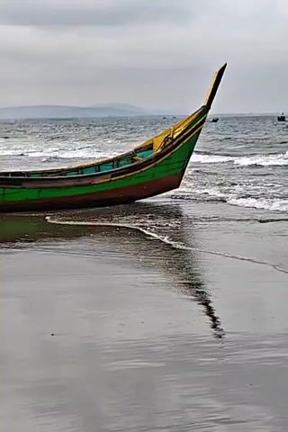 A fishing boat is pulled up on a sandy beach. The boat is green and yellow. The ocean waves are coming in. The sky is cloudy and overcast. The water is reflecting the boat and the sky.