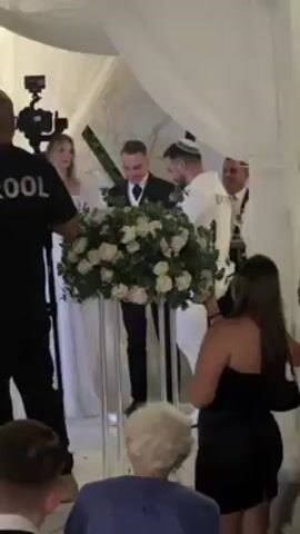 The video shows a wedding celebration under a white canopy. A man in a black suit is standing at a podium adorned with white roses, and appears to be making a speech or announcement.  A bride in a white dress stands nearby, with another man in a white shirt to her right.  People, mostly in formal attire, are gathered around, some clapping and raising their hands in celebration.  A person wearing a shirt with the word "KROOL" on the back is visible on the left, standing near a camera. The setting appears to be an indoor space, likely a venue for special events.