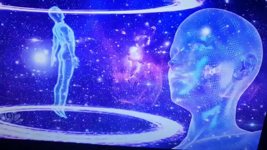 The video shows an animated digital rendering of a human figure in a cosmic or celestial environment. The figure is translucent and appears to be made of glowing blue lines forming a wireframe of a human body. Around the figure, there are swirling rings of light, some appearing pinkish and others white. In the background, there is a deep blue expanse filled with stars and nebulae. To the right of the frame, a large, translucent, wireframe head is visible, also composed of blue lines, facing left towards the human figure. In the lower left corner, there is a logo featuring a lotus flower with the word "VIBRATION" and "THERAPIES" underneath it. The scene transitions slightly, showing two large, translucent wireframe hands reaching up from below, framing the human figure. The overall aesthetic is futuristic and spiritual, suggesting themes of energy, consciousness, or healing.