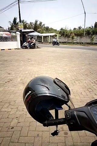The video opens with a close-up view of a black Honda motorcycle helmet hanging from the handlebars of a parked scooter. The scooter's dashboard and handlebars are visible in the foreground. The camera then pans slightly to reveal a paved area made of brick-like stones. In the background, a road is visible, with a few trees and vegetation lining the sides. Across the road, there are several small buildings or stalls, one of which has a white and red sign. Two people are on a scooter near one of these stalls, appearing to be interacting. Further down the road, another scooter with a rider is visible in motion. The sky is bright and clear, indicating daytime. The overall scene appears to be an outdoor setting, possibly a roadside stop or a small market area.