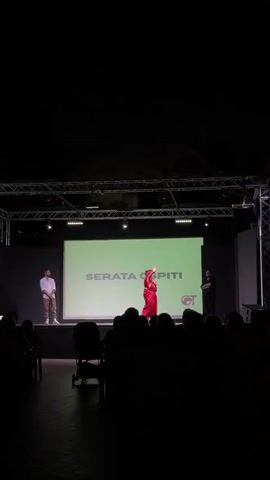 A short clip shows a stage setup at night. A large screen in the background displays "SERATA OSPITI" in white text on a light green background, with a circular logo to the right. Three individuals are on stage. On the left, a person stands facing the screen, appearing to be a presenter. In the center, a person in a bright red outfit is performing or dancing, with their arms outstretched and head tilted back. To the right of the performer, another person stands facing the audience, seemingly a host or presenter. In the foreground, the silhouettes of an audience are visible, seated and facing the stage. The lighting is focused on the stage, with the background dark.