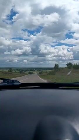A first-person view from inside a vehicle shows a road stretching into the distance under a dramatic sky. The road is paved and has a white line marking the center. On either side of the road, there is green grass and some trees. In the background, a landscape of rolling hills and trees can be seen. The sky is filled with large, fluffy white clouds against a blue background, suggesting it is daytime and the weather is partly cloudy, possibly with a chance of rain. The overall mood is one of travel and openness, with the vast sky and open road creating a sense of journey. The time of day appears to be daytime, likely late morning or afternoon, given the brightness of the light. The location is indicated as Agapovka, Russia, which aligns with the rural, pastoral scenery. There is no visible text in the media.
