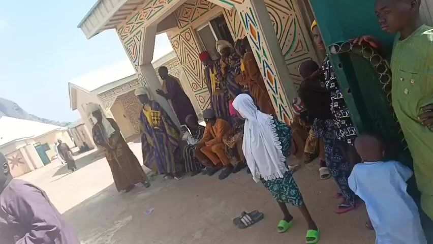 The video depicts a group of people gathered in a courtyard in front of a building with decorative geometric patterns. Several men are wearing traditional robes and hats.  There's a small group of musicians with a trumpet and drums. Some people are standing and observing, while others are seated. There is a mountain in the background. The video appears to be shot in a sunny, outdoor environment.