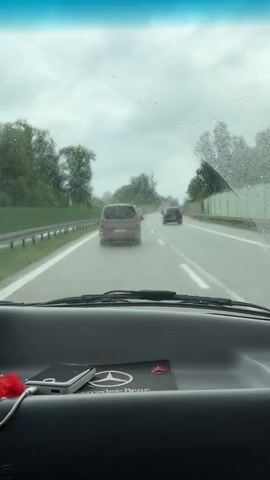 The video shows the interior of a car driving on a highway in rainy conditions. The windshield wipers are in use. The dashboard includes a Mercedes-Benz branded item, and a smartphone connected to a red, fuzzy pompom. The side mirror shows a reflection of a person in the back seat taking a photo or video of themselves with a smartphone. The highway has other vehicles on it and has a barrier on the right. Trees are visible beyond the barrier.