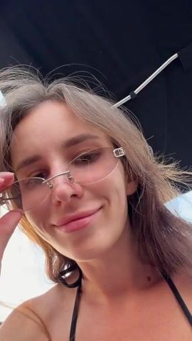 A woman with brown hair and sunglasses is on a boat. She is wearing a black bikini top and smiles at the camera, sticking out her tongue. The sky is visible in the background.