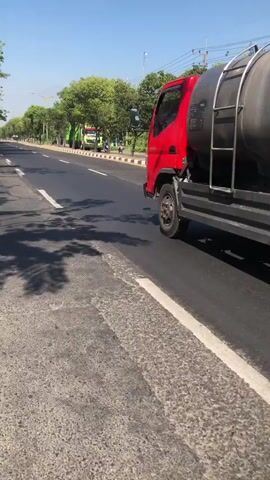 A red and black tank truck drives down a paved road lined with trees and a sidewalk. Another green truck follows. A person on a motorcycle with a white box on the back rides behind them. The sun shines, casting shadows from the trees onto the road.