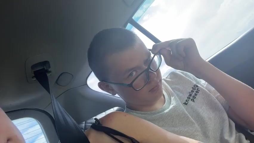 A person with short hair and glasses is sitting in the back seat of a car. They are adjusting their glasses and looking at the camera with a playful expression. The sun is shining through the window, creating a bright backlight. A seatbelt is visible on the left side of the frame. The person is wearing a light-colored t-shirt with text on it.