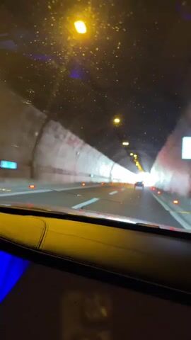 A first-person perspective video shows a car driving through a tunnel. The tunnel is lit by yellow overhead lights and has a bright light at the end. The road ahead is visible, with lane markings and orange markers on the sides. Another car is visible ahead, driving in the same direction. The interior of the car in which the video is being filmed is partially visible in the foreground, showing a yellow quilted dashboard and blue ambient lighting. The video is static, with no visible movement other than the progress through the tunnel.