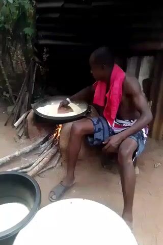 A shirtless man with a pink towel around his neck is seated on a stool and stirring a white liquid in a large pan over an open fire.  The man is wearing blue and white patterned shorts and gray sandals.  The fire is fueled by pieces of wood stacked beneath the pan.  Two buckets, one black and one white, containing the same white liquid as the pan, are positioned in front of the man. Behind the man is a crude wooden structure with a corrugated metal roof. The ground is dirt. Green foliage is visible in the background. The man uses a brown, round, flat object to stir the liquid.