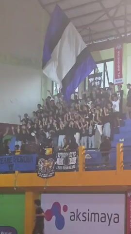 A crowd of people, likely spectators at a sporting event, are gathered in tiered seating. A large flag with horizontal stripes of purple, white, and black is prominently displayed, held aloft by some individuals in the crowd. Several banners are visible in the foreground, one reading "SPEROSA BOYS." The environment appears to be an indoor venue, possibly a gymnasium or arena, with green and yellow walls and windows in the background. The lighting suggests it is daytime. Some individuals are raising their hands, possibly cheering or reacting to an event. The overall atmosphere seems to be one of excitement and support for a team or group.