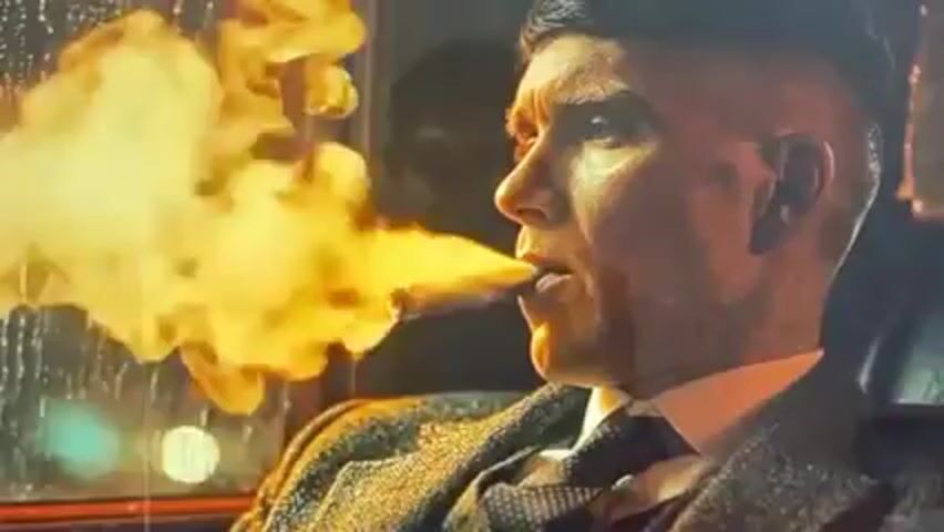 A man with dark, slicked-back hair and a short haircut is smoking a cigar in a car at night. He is wearing a patterned tweed jacket, a white shirt, and a dark tie. The cigar emits a large cloud of smoke that obscures part of his face. Rain streaks across the window next to him, and a blurry light is visible outside.