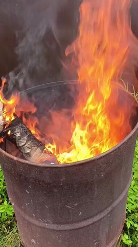 The video shows a close-up shot of a large, rusty metal barrel with a fire burning inside. The flames are bright orange and yellow, with some red and white hues, and they are reaching upwards out of the barrel. There is a piece of wood burning on the left side of the barrel. Smoke is visible rising from the fire and billowing to the left. The barrel is sitting outdoors, with green foliage visible around its base and in the background. The camera is positioned at a slight downward angle, looking into the barrel. The lighting suggests it is daytime.