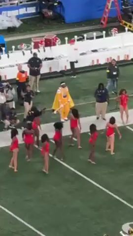 The video shows a group of approximately ten dancers in matching red leotards performing on a football field. They are arranged in rows and columns and execute synchronized moves. Surrounding them is a group of people including photographers, security personnel, and a person in a bright yellow jumpsuit and cape. The field is marked with white lines and there are various pieces of equipment and structures visible in the background.