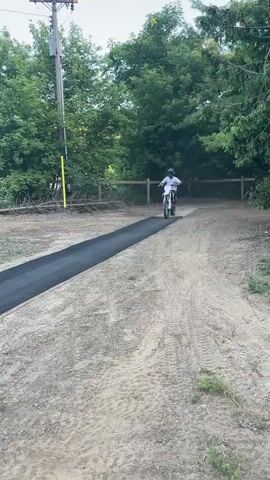 A person on a dirt bike rides down a dirt path towards a black ramp. The rider, wearing a helmet and protective gear, speeds up and launches off the ramp into the air. The rider and bike are captured mid-air, with trees and an inflatable airbag structure in the background. The rider appears to be performing a jump.
