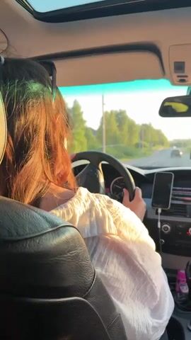 The video shows the interior of a car from the perspective of the back seat, looking forward. A person with long, dark, wavy hair, wearing a white shirt, is driving the car, holding the steering wheel with both hands. Sunlight illuminates their hair and shirt. The car's dashboard is visible, including a phone mounted in a holder near the center console, connected to a charging cable. Through the windshield, a road is visible, flanked by green trees and fields under a bright sky. The car appears to be moving.