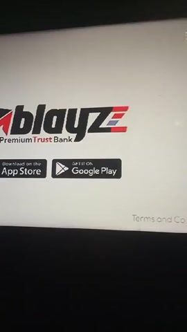 The video shows a close-up of a television screen displaying the logos for Blayz by Premium Trust Bank, followed by the Premium Trust Bank logo. The screen also shows icons for downloading the Blayz app from the Apple App Store and Google Play. Text on the screen states "Nigeria's fastest growing bank". The camera is stable. The video appears to be taken indoors, as indicated by the reflections on the screen. The background is dark.