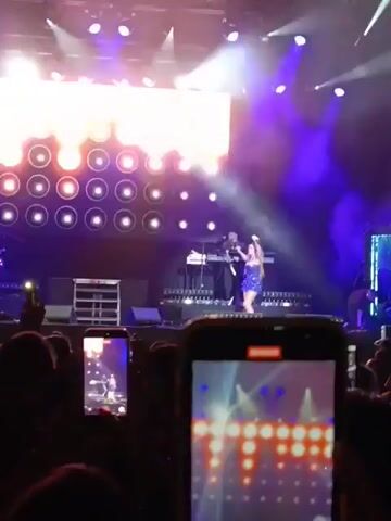 A woman in a short blue dress is performing on a stage in front of a large crowd. She is singing into a microphone, gesturing, and moving around the stage. The stage has bright lights and a large video screen in the background displaying what appear to be the same patterns as on the smartphones recording the performance.  There are musicians visible behind her, and the crowd is recording the performance with their phones. The recording is shaky and primarily focused on one of the phones.