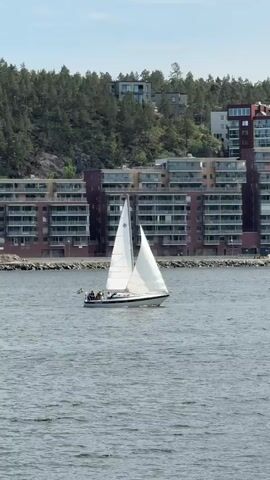 A sailboat with white sails and a Swedish flag is sailing across a body of water. The sailboat has several people on board. In the background, there are apartment buildings and trees on a hill. The water is a dark blue-grey color, and the sky is light blue with some white clouds. The sailboat is moving from right to left in the frame.