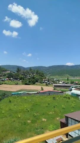 The video shows a person standing on a balcony, looking out at a village nestled in a valley. The person is wearing a yellow hat, a blue t-shirt, and khaki shorts, and has a black backpack on. The village has houses with colorful roofs, and is surrounded by green fields and rolling hills covered in trees. The sky is clear and blue with a few white clouds. The balcony has a brown railing with yellow metal supports. The camera pans slightly to the left, showing more of the village and the surrounding landscape. The person occasionally shifts their weight and adjusts their grip on the railing, but otherwise remains stationary, observing the view. The video appears to be shot in natural daylight.