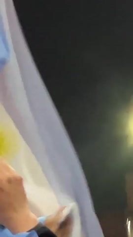 A person's hand with a white manicure is briefly visible in the lower left corner of the frame, touching a white fabric that billows upwards. The scene then shifts to a group of people at night, holding and waving an Argentinian flag. The flag, with its characteristic light blue and white stripes and a sun emblem, is prominently displayed. In the background, a dark sky is visible, with a few distant lights and some trees. A person in the background appears to be wearing a jersey with the number 10. Another person is partially visible behind the flag, wearing a black cap with a white emblem. The overall atmosphere suggests a celebration or event related to Argentina.