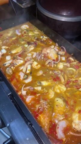 The video shows a large rectangular pan on a stovetop, filled with a stew-like dish. The dish contains a variety of seafood, including shrimp, mussels, and squid, along with pieces of chicken and red bell peppers. The stew is simmering and producing steam.  The scene is close-up, focusing on the dish, with a glimpse of a stainless steel pot above.